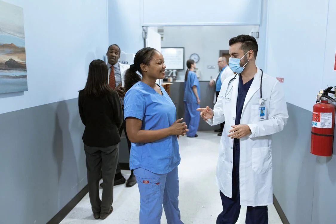 A nurse in blue scrubs speaks with a doctor in a white coat in a hospital corridor, with colleagues in the background.